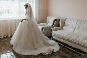 le la mariée dans une blanc mariage robe. content magnifique Jeune femme dans blanc traditionnel mariage robe dans sa chambre. grand les fenêtres et lumière des murs. photo