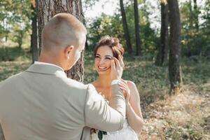 mariage photo dans la nature. le la mariée et jeune marié sont permanent près une arbre. le la mariée regards à le marié, et il doucement corrige sa boucles. réunion dans le forêt.