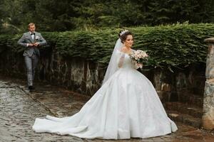 une Beau jeune marié et une charmant la mariée sont en marchant ensemble près le pierre clôtures dans le parc. luxueux robe avec une longue train. jeune marié de derrière photo