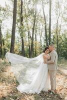 mariage photo. le la mariée et jeune marié sont permanent dans le forêt étreindre, le jeune marié tendrement baisers le sien bien-aimée sur le front. une voile cette chutes magnifiquement dans le air. couple dans l'amour. été lumière. photo