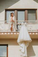 blanc mariage robe pendaison sur balcon balustrade, large coup de maison dans de face et fille sur balcon. mariage détails, moderne mariage robe avec longue former, longue manches et ouvert retour photo