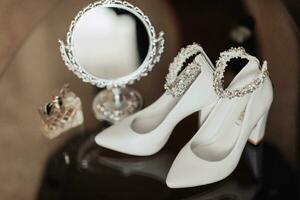 photo de détails à le mariage. blanc élégant des chaussures avec décoration sur le pied, une magnifique miroir avec une argent Cadre et parfum supporter sur une verre rester. ombres et lumière.