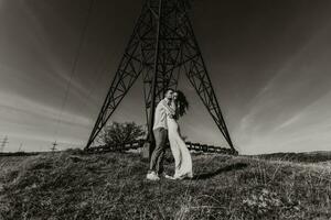 élégant modèle couple dans le montagnes dans été. une Jeune garçon et fille dans une blanc soie robe sont permanent près grand les structures de Puissance lignes. noir et blanc contraste photo