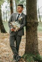 une élégant jeune marié avec une bouquet de fleurs dans le sien mains est permanent près une arbre dans le forêt. plein longueur photo