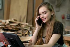 une Jeune femme est formation à être une Charpentier dans le atelier. elle travaux avec une portable ordinateur dans une bois atelier. femelle Charpentier contact les clients par téléphone intelligent. pme ordres, Commencez et petit photo