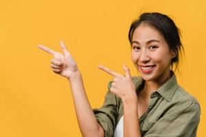 portrait d'une jeune femme asiatique souriante avec une expression joyeuse, montre quelque chose d'étonnant dans un espace vide dans un tissu décontracté et regardant la caméra isolée sur fond jaune. concept d'expression faciale. photo