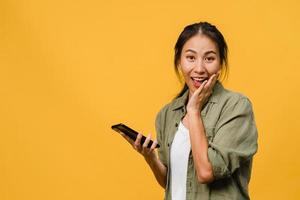 surpris une jeune femme asiatique utilisant un téléphone portable avec une expression positive, un large sourire, vêtue de vêtements décontractés et regardant la caméra sur fond jaune. heureuse adorable femme heureuse se réjouit du succès. photo