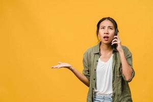 une jeune femme asiatique parle par téléphone avec une expression négative, des cris excités, des cris émotionnels en colère dans un tissu décontracté et se tient isolée sur fond jaune avec un espace de copie vierge. concept d'expression faciale. photo