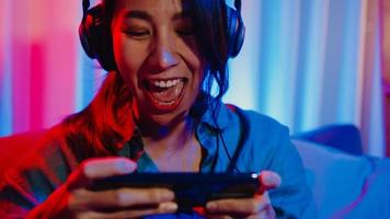 une joyeuse joueuse asiatique porte un jeu vidéo de compétition de casque en ligne avec une conversation excitée par smartphone avec un ami assis sur un canapé dans un salon aux néons colorés à la maison, concept d'activité de quarantaine à domicile.