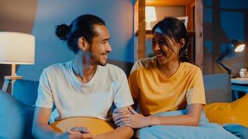 heureux jeune couple asiatique homme et femme regardant la caméra sourire et joyeux lors d'un appel vidéo en ligne la nuit dans le salon à la maison, rester à la maison en quarantaine, vie conjugale, concept de distanciation sociale. photo