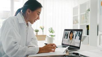 jeune femme médecin asiatique en uniforme médical blanc utilisant un ordinateur portable parlant par vidéoconférence avec un médecin senior au bureau d'une clinique de santé ou d'un hôpital. distanciation sociale, quarantaine pour le virus corona.