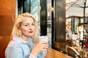 portrait de une Jeune femme en buvant café ou thé dans une café. le fille est habillé dans une bleu chemise, blond. regards directement dans le caméra. photo