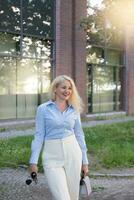 une réussi femme d'affaires dans une bleu chemise et blanc pantalon est permanent à l'extérieur le bureau. une chaud ensoleillé journée. photo