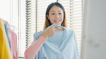 belle dame asiatique séduisante choisissant des vêtements sur un portemanteau s'habillant en se regardant dans un miroir dans la chambre de la maison. fille pense quoi porter chemise décontractée. les femmes de style de vie se détendent à la maison concept. photo