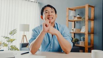 un jeune homme d'affaires asiatique utilisant un ordinateur portable parle à ses collègues du plan lors d'une réunion par appel vidéo tout en travaillant à domicile dans le salon. auto-isolement, distanciation sociale, quarantaine pour le virus corona.