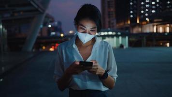 jeune femme d'affaires asiatique en vêtements de mode portant un masque facial à l'aide d'un téléphone intelligent en tapant un message texte tout en se tenant à l'extérieur dans une ville urbaine la nuit. distanciation sociale pour empêcher la propagation du concept covid-19.