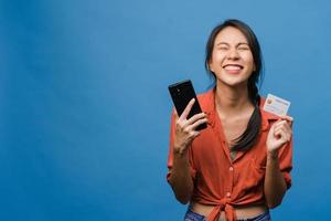 jeune femme asiatique utilisant un téléphone et une carte bancaire de crédit avec une expression positive, sourit largement, vêtue de vêtements décontractés et se tient isolée sur fond bleu. heureuse adorable femme heureuse se réjouit du succès. photo