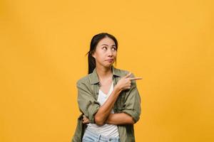 une jeune femme asiatique montre quelque chose d'étonnant dans un espace vide avec une expression négative, des cris excités, des pleurs émotionnels en colère dans des vêtements décontractés isolés sur fond jaune. concept d'expression faciale. photo
