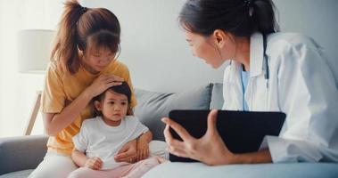 jeune médecin pédiatre asiatique et petite fille patiente utilisant une tablette numérique partageant de bonnes nouvelles sur les tests de santé avec une maman heureuse s'asseoir sur un canapé dans la maison. assurance médicale, rendre visite au patient à domicile concept.