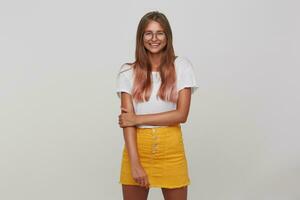 bien à la recherche positif Jeune longue aux cheveux blond femme souriant Heureusement tandis que permanent plus de blanc arrière-plan, habillé dans blanc de base T-shirt et Jaune jupe photo