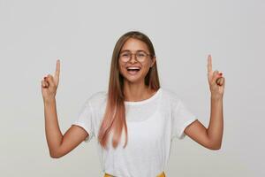 content Jeune charmant blond Dame avec longue cheveux montrant vers le haut avec élevé index et rire gaiement, portant blanc de base T-shirt tandis que permanent plus de blanc Contexte photo