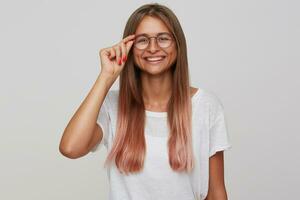 content Jeune magnifique blond femme en portant sa des lunettes tandis que à la recherche positivement à caméra et souriant gaiement, étant dans agréable ambiance tandis que permanent plus de blanc Contexte photo