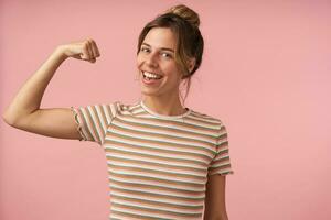 studio photo de charmant Jeune brunette femelle habillé dans beige rayé T-shirt élevage main tandis que démontrant sa Puissance et souriant volontier à caméra, isolé plus de rose Contexte