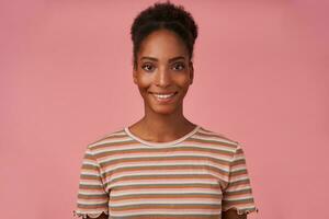 portrait de Jeune magnifique marron aux cheveux frisé Dame avec foncé peau montrant sa blanc parfait les dents tandis que à la recherche Heureusement à caméra, isolé plus de rose Contexte photo