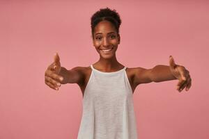 de bonne humeur Jeune jolie marron aux cheveux frisé femme avec foncé peau souriant Heureusement à caméra tandis que permanent plus de rose Contexte avec ouvert bras, Aller à donner étreinte à Quelqu'un photo