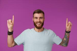 joyeux Jeune non rasé tatoué homme avec court la Coupe de cheveux portant bleu T-shirt tandis que permanent plus de violet arrière-plan, à la recherche à caméra avec large content sourire et élevage en haut le sien indice les doigts photo