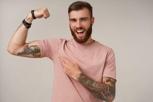 joyeux Jeune barbu brunette Masculin avec branché la Coupe de cheveux portant beige T-shirt posant plus de blanc arrière-plan, montrant sur le sien élevé main et sentiment fier à propos fort muscles photo