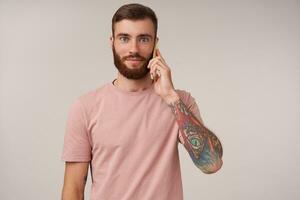 charmant Jeune aux yeux bleus tatoué brunette homme avec tatouages fabrication appel avec le sien téléphone intelligent et à la recherche à caméra avec positif affronter, portant beige T-shirt tandis que permanent plus de blanc Contexte photo