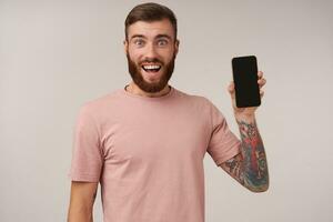 content aux yeux bleus jolie barbu Masculin avec tatouages portant beige T-shirt tandis que posant plus de blanc arrière-plan, souriant joyeusement à caméra et élevage main avec téléphone intelligent dans il photo