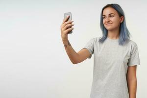 intérieur photo de Jeune court aux cheveux positif tatoué femelle en gardant mobile téléphone dans élevé main et souriant agréablement à caméra, isolé plus de blanc Contexte