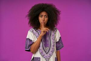 sérieux Jeune foncé écorché Dame avec décontractée coiffure posant plus de violet arrière-plan, renfrogné et élevage indice doigt dans silence geste, portant blanc à motifs chemise photo