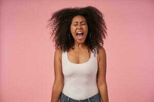 stressé Jeune foncé écorché femme avec frisé cheveux posant plus de rose Contexte avec mains bas, renfrogné sa visage avec fermé yeux et en hurlant bruyant avec large bouche ouvert photo