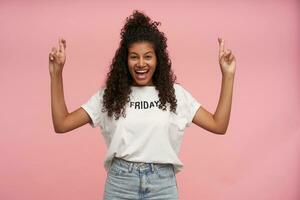 joyeux attrayant Jeune frisé brunette Dame avec foncé peau traversée les doigts pour bien la chance et à la recherche gaiement à caméra avec vaste sincère sourire, porter blanc T-shirt et jeans plus de rose Contexte photo