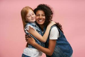 de bonne humeur attrayant Jeune foncé écorché brunette femme avec longue frisé cheveux souriant Heureusement à caméra et câlins mignonne positif roux femelle enfant, isolé plus de rose Contexte photo