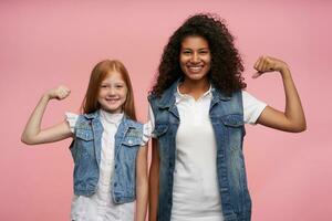 content charmant longue aux cheveux Jeune les filles dans décontractée Regardez élevage mains et montrant leur pouvoir, à la recherche gaiement à caméra avec charmant sourires, permanent contre rose Contexte photo