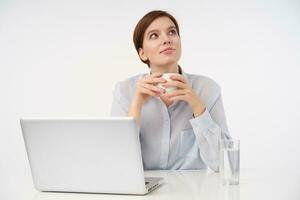 agréable à la recherche Jeune court aux cheveux brunette Dame avec décontractée coiffure souriant positivement tandis que en pensant à propos quelque chose positif et en buvant tasse de café, isolé plus de blanc Bureau Contexte photo