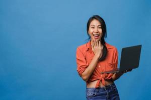surpris une jeune femme asiatique utilisant un ordinateur portable avec une expression positive, un large sourire, vêtue de vêtements décontractés et regardant la caméra sur fond bleu. heureuse adorable femme heureuse se réjouit du succès.