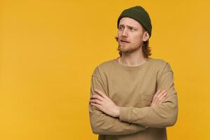 portrait de beau, adulte Masculin avec blond cheveux et barbe. portant vert bonnet et beige chandail. garde bras franchi. en train de regarder à le la gauche à copie espace, isoler plus de Jaune Contexte photo