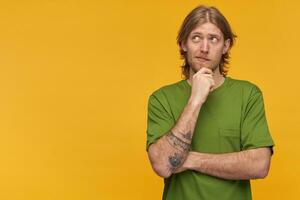 portrait de rêver Masculin avec blond coiffure et barbe. portant vert T-shirt. a tatouage. émouvant le sien menton. en train de regarder pensivement à le la gauche à copie espace, isolé plus de Jaune Contexte photo