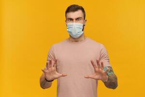 effrayé Jeune tatoué barbu homme dans rose t chemise et hygiénique masque à prévenir infection avec mains dans de face de lui-même et défendre de menace plus de Jaune Contexte photo