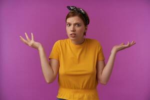 portrait de attrayant Jeune marron regardé européen femme avec Naturel maquillage émotion confus devine avec bras de côté portant élégant Jaune T-shirt et noir bandana posant contre violet Contexte photo