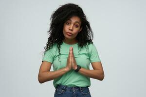 malheureux désespéré africain américain Jeune femme avec longue frisé cheveux dans menthe T-shirt garde mains dans prier position et demander pour Aidez-moi isolé plus de blanc Contexte photo