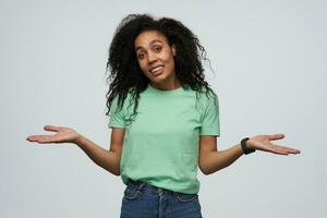 souriant confus africain américain Jeune femme avec frisé longue cheveux dans menthe T-shirt permanent et en portant fond sur deux paumes isolé plus de blanc Contexte photo