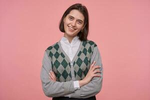 intérieur coup de Jeune brunette femelle porte élégant gris arrêtez-vous souriant tandis que mettant en vedette dans caméra et franchi sa mains sur poitrine. isolé plus de rose Contexte. photo