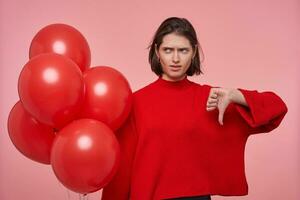 intérieur portrait de Jeune brunette femme, porte brillant rouge chandail orage des ballons et spectacles les pouces vers le bas signe, tombe malheureux, photo