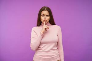 sérieux Jeune jolie marron aux cheveux femme avec Naturel maquillage renfrogné sa les sourcils tandis que à la recherche gravement à caméra, élevage main avec silence geste tandis que posant plus de violet Contexte photo